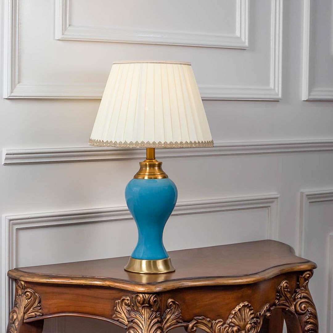 Blue ceramic table lamp turned on with pleated fabric shade and brass detailing displayed on white background.