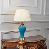 Blue ceramic table lamp turned on with pleated fabric shade and brass detailing displayed on white background.