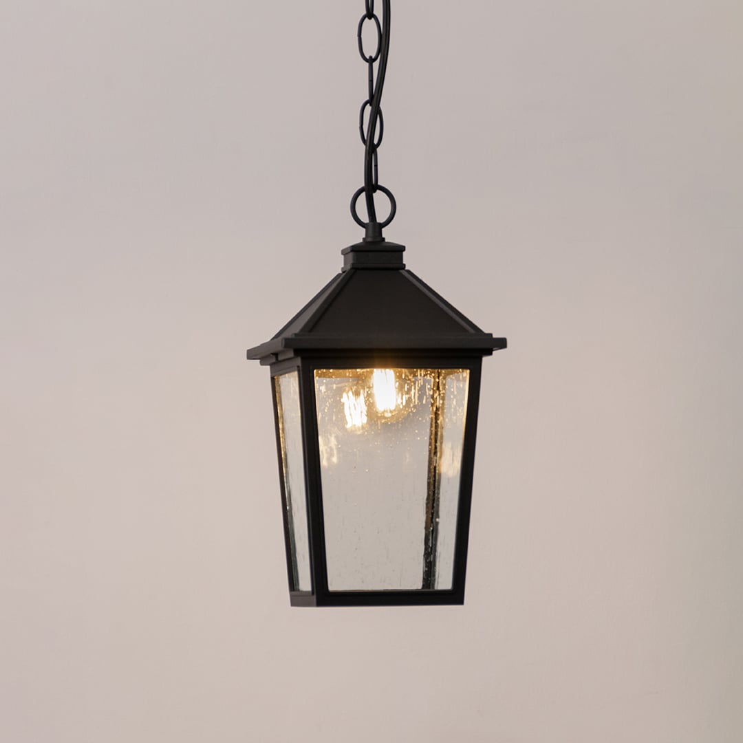 Black outdoor lantern with the light switched on displayed on a white background showing a warm glow through textured clear glass.