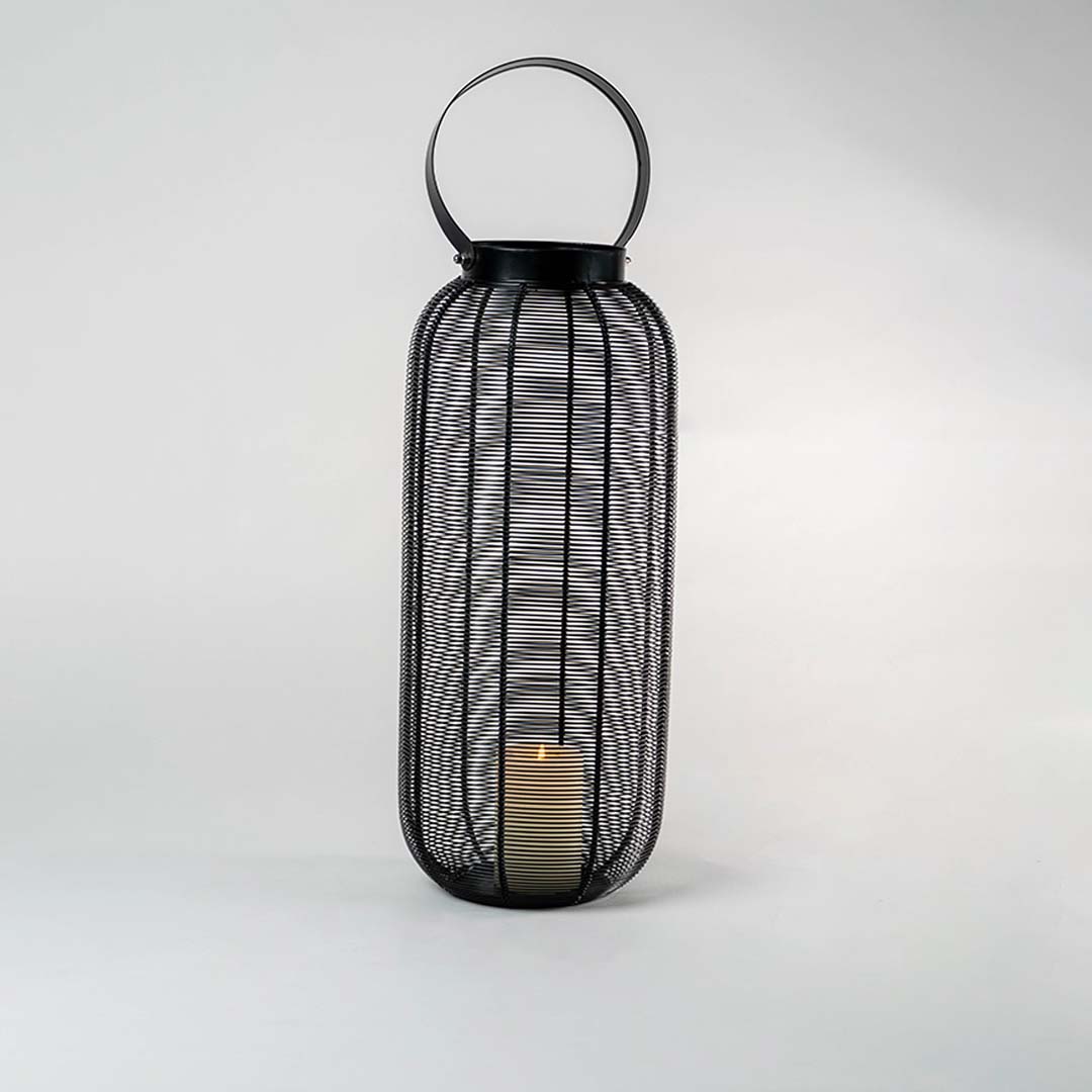 Illuminated tall black wire lantern isolated on white background showing warm candle glow casting intricate shadows through mesh frame.