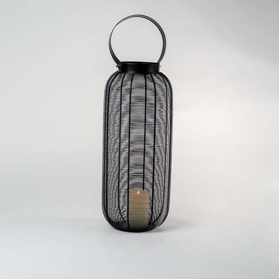 Illuminated tall black wire lantern isolated on white background showing warm candle glow casting intricate shadows through mesh frame.