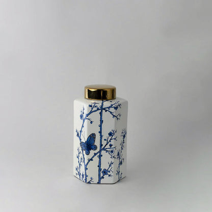 White ceramic jar with blue floral pattern and gold lid placed on white surface against neutral background showcasing elegant classic home decor styling.