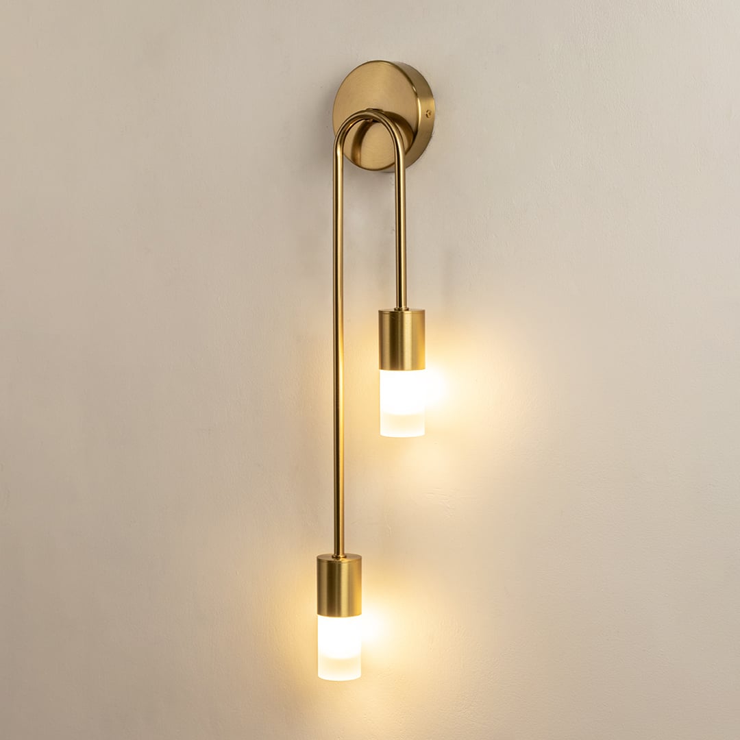 A gold metal dual wall sconce with frosted glass cylinder shades shown with lights on against a white background highlighting its warm soft illumination.