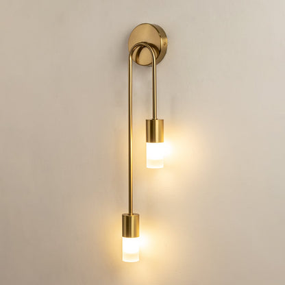 A gold metal dual wall sconce with frosted glass cylinder shades shown with lights on against a white background highlighting its warm soft illumination.