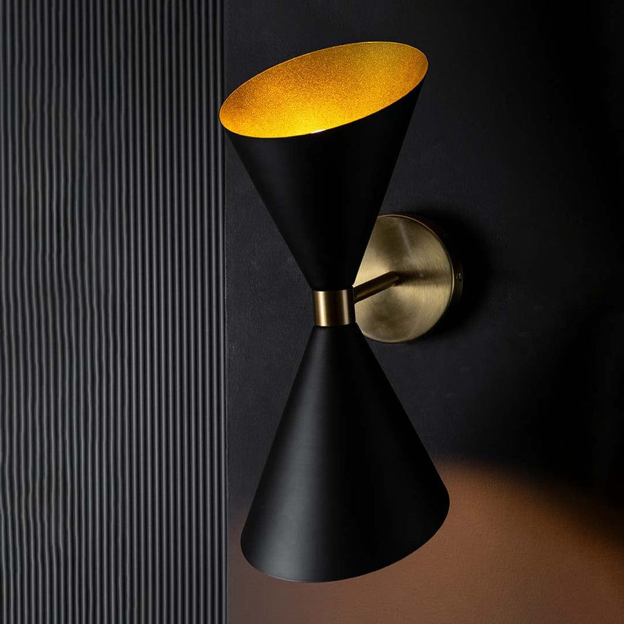 Black and gold double shade wall light shown switched on against a dark background with warm light casting an elegant glow above and below the fixture.