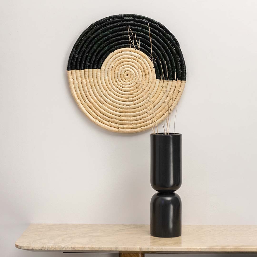 Round black and beige woven wall hanging styled with black ceramic vase for elegant home wall decor.