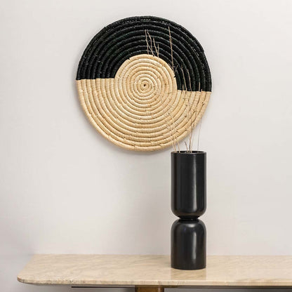 Round black and beige woven wall hanging styled with black ceramic vase for elegant home wall decor.