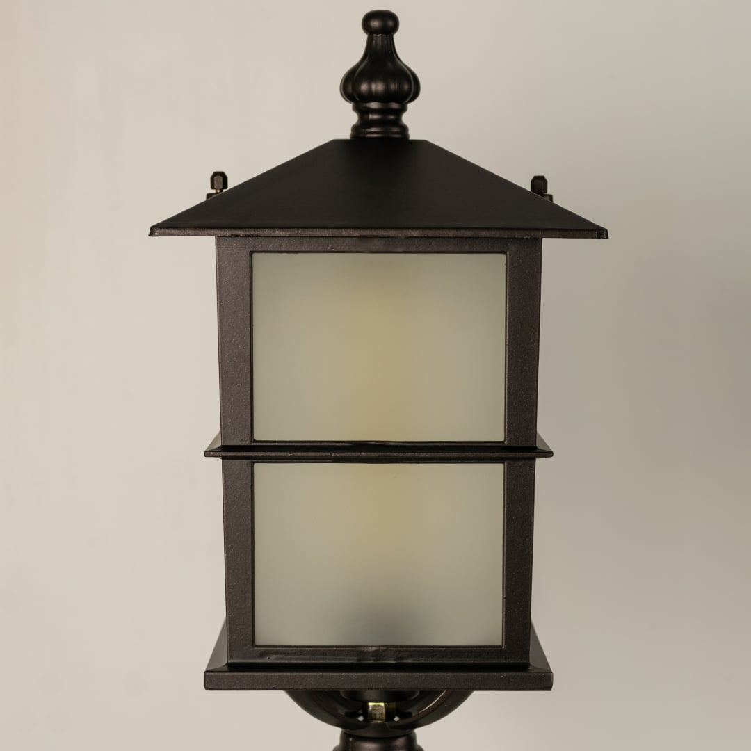 Closeup of a black outdoor pillar lamp showing glowing frosted glass and refined lantern edges designed for soft ambient garden and pathway lighting.