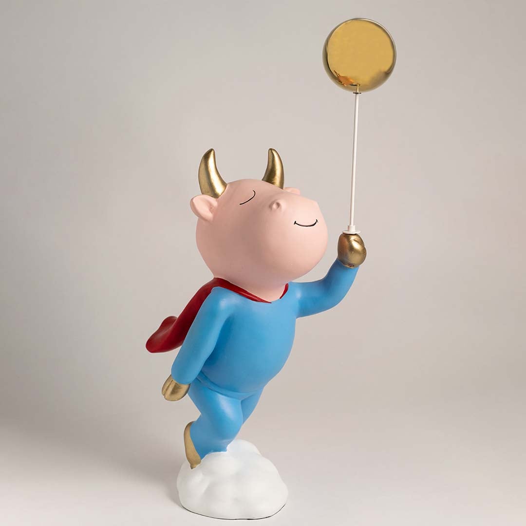 A playful decorative figurine of a smiling character in a blue outfit with gold details and a red cape standing on a cloud and holding a golden balloon on a stick.