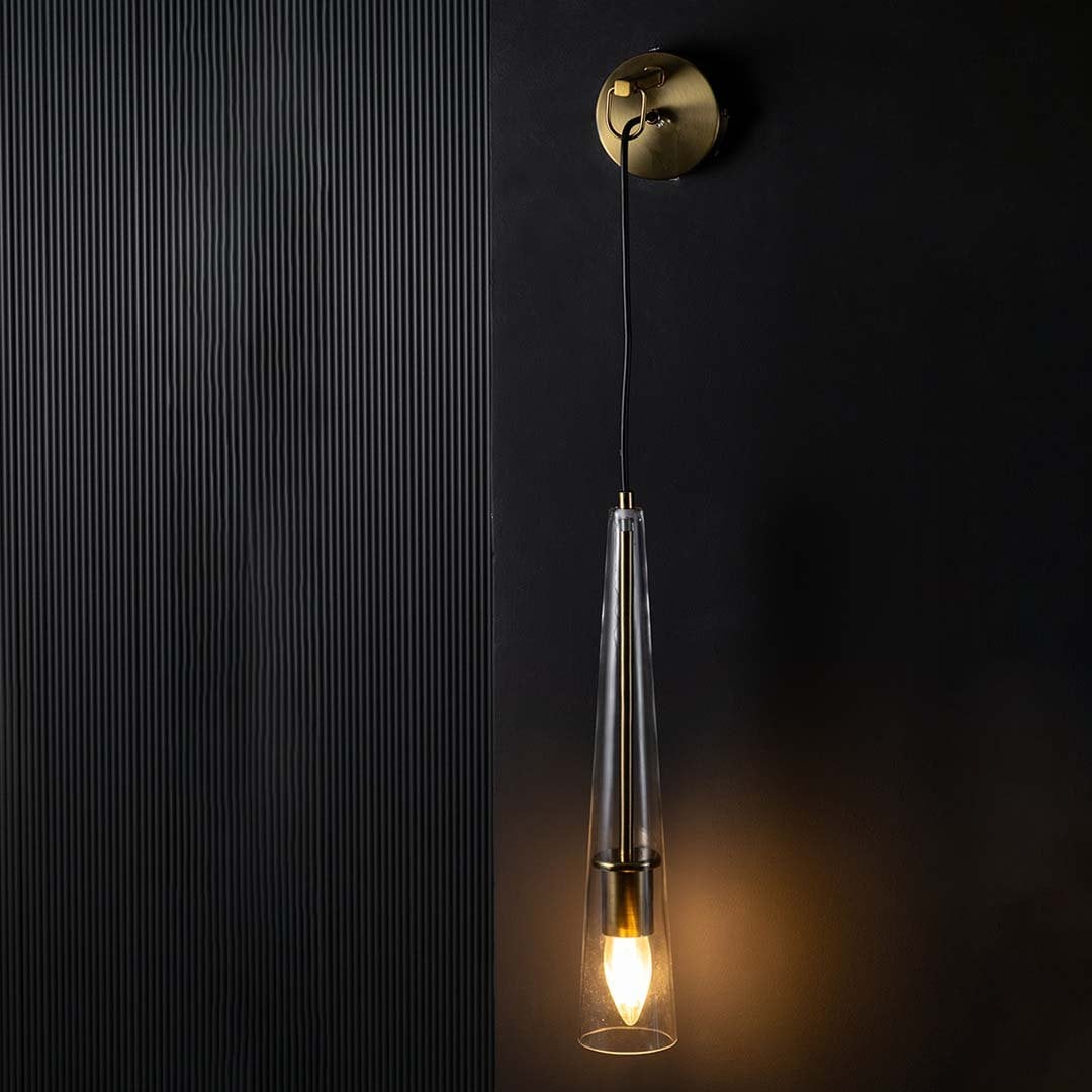 Glowing brass wall light fixture with hanging tapered glass shade set against black background emphasizing dramatic ambiance and design.
