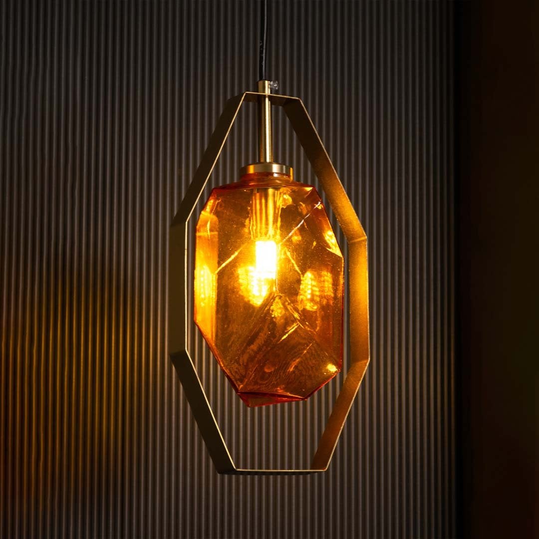 Glowing amber rock glass pendant light with gold frame set against dark background highlighting geometric shape and warm luxury ambiance.
