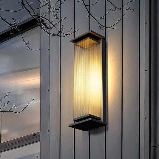 Modern black rectangular outdoor wall light with vertical ribbed glass illuminated on grey siding near tree branches at dusk.