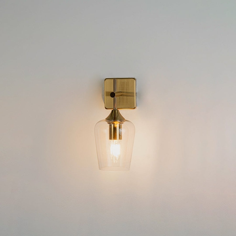 Clear glass wall light with gold metal base shown on a white background with the light on displaying a warm glow for modern bedroom hallway or living room decor.