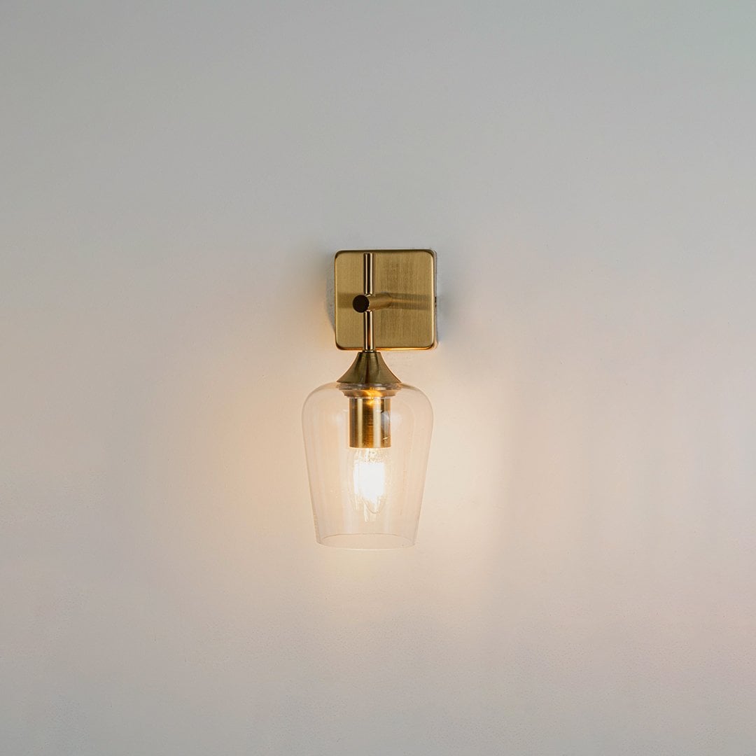 Clear glass wall light with gold metal base shown on a white background with the light on displaying a warm glow for modern bedroom hallway or living room decor.