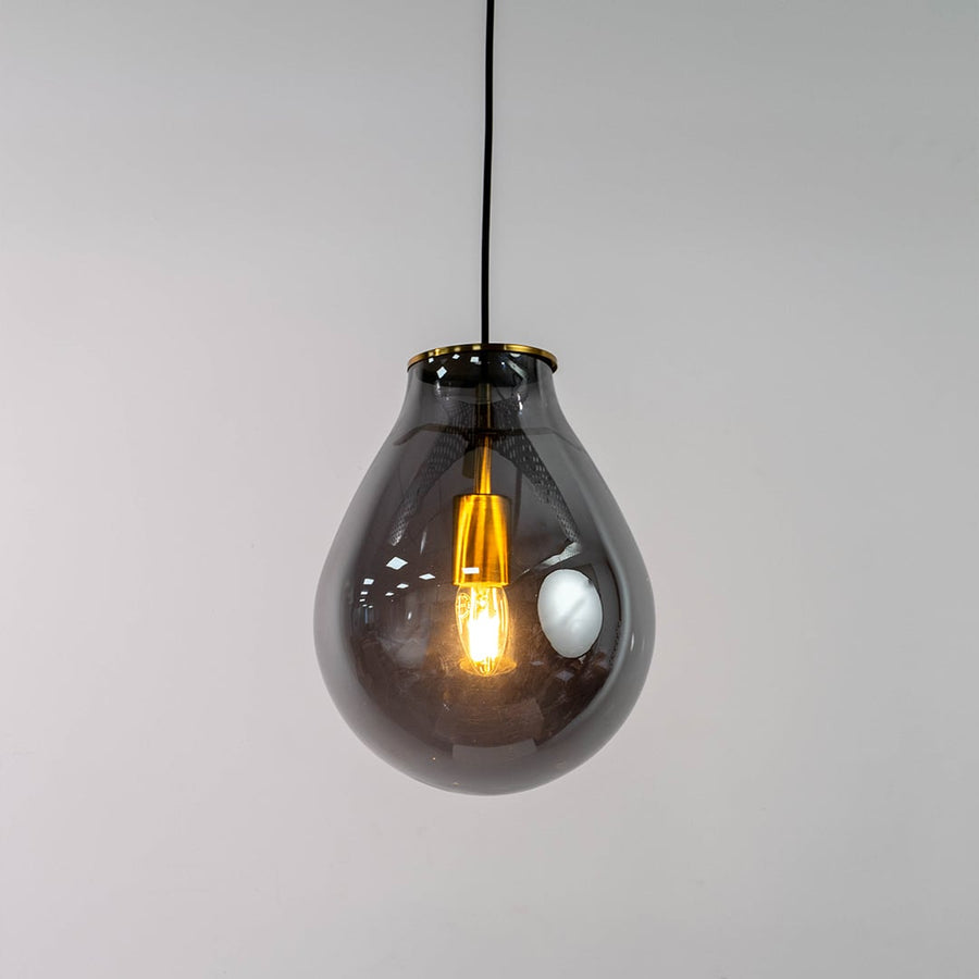 A smoked grey glass pendant light with lights on against a clean white background showing its teardrop shaped glass shade and warm glowing filament.