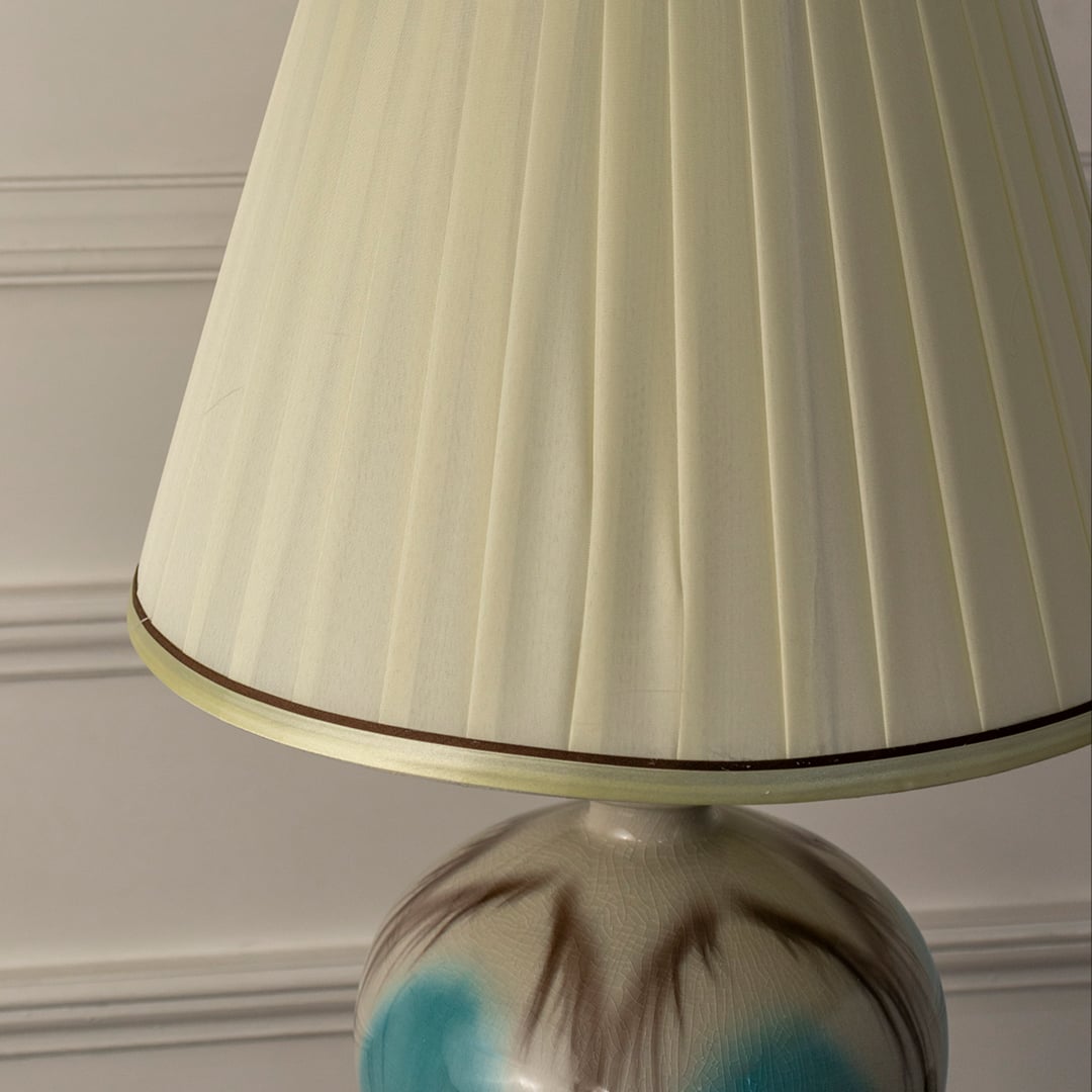 Closeup of blue ceramic table lamp featuring glossy gradient finish gold detailing and soft pleated white lampshade.