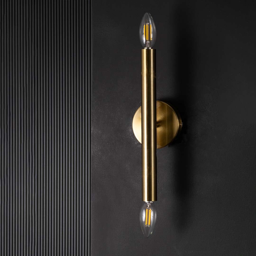 Gold metal vertical wall light with bulbs off against a black backdrop showing its slim cylindrical frame and modern minimal design.