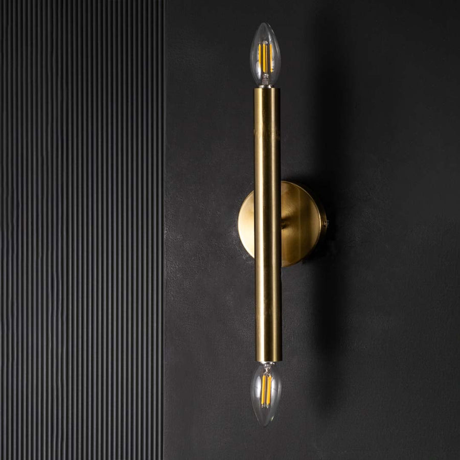 Gold metal vertical wall light with bulbs off against a black backdrop showing its slim cylindrical frame and modern minimal design.