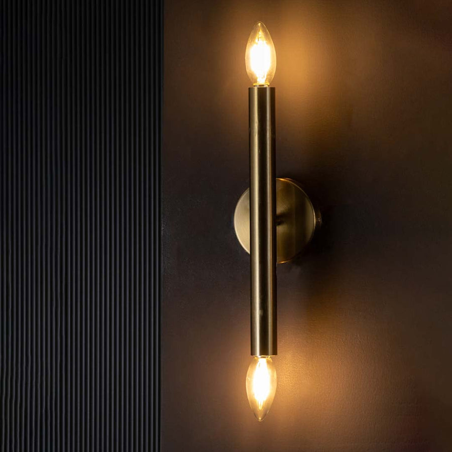 Gold double ended wall light glowing warmly on a dark background showcasing its sleek metal tube and bright ambient vertical lighting effect.