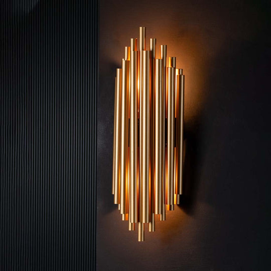 A gold metal wall light with lights on glowing softly against a dark black background emphasizing the sleek vertical metal tube design.
