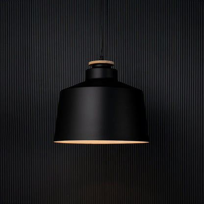 A modern black pendant lamp shown on a black background with the light on creating a soft warm glow that highlights the shape and contrasts against the dark surface.