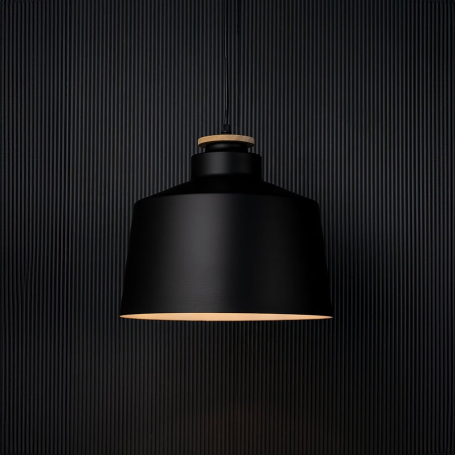 A modern black pendant lamp shown on a black background with the light on creating a soft warm glow that highlights the shape and contrasts against the dark surface.
