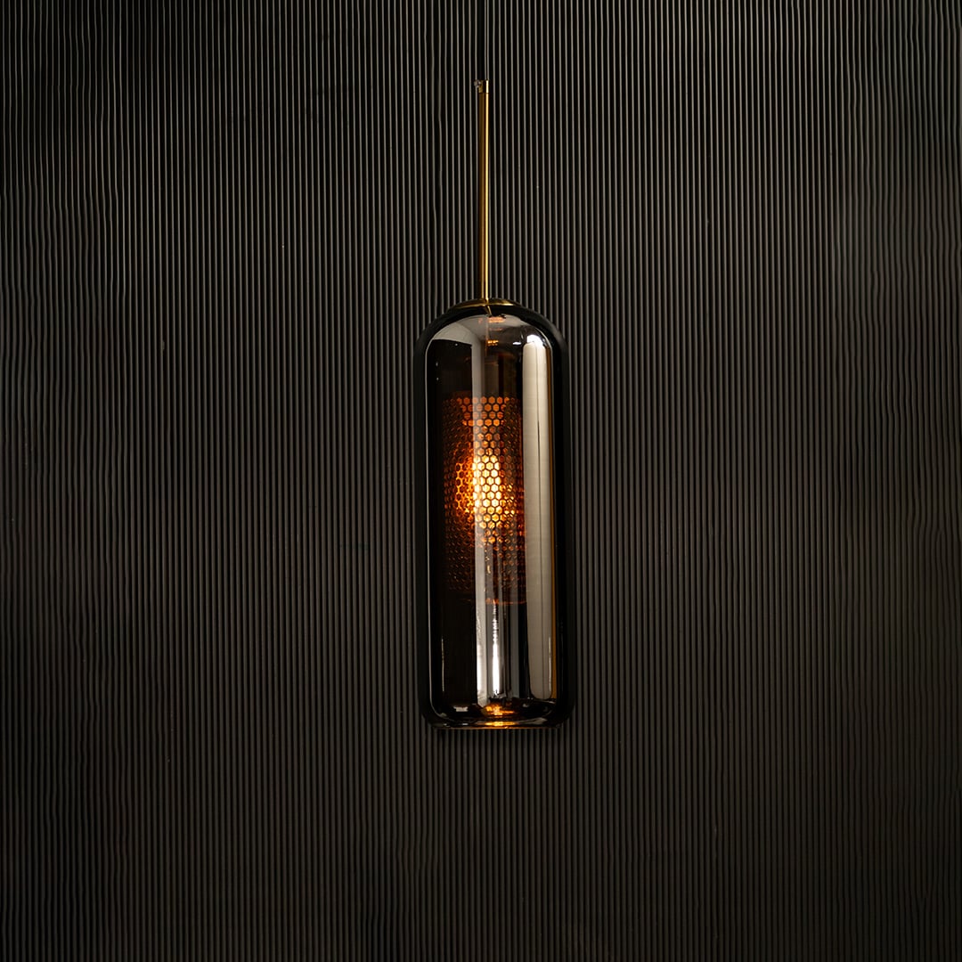 Tall smoked grey glass pendant with gold fitting and warm glowing diffuser highlighted on a dark background creating a dramatic stylish lighting accent.