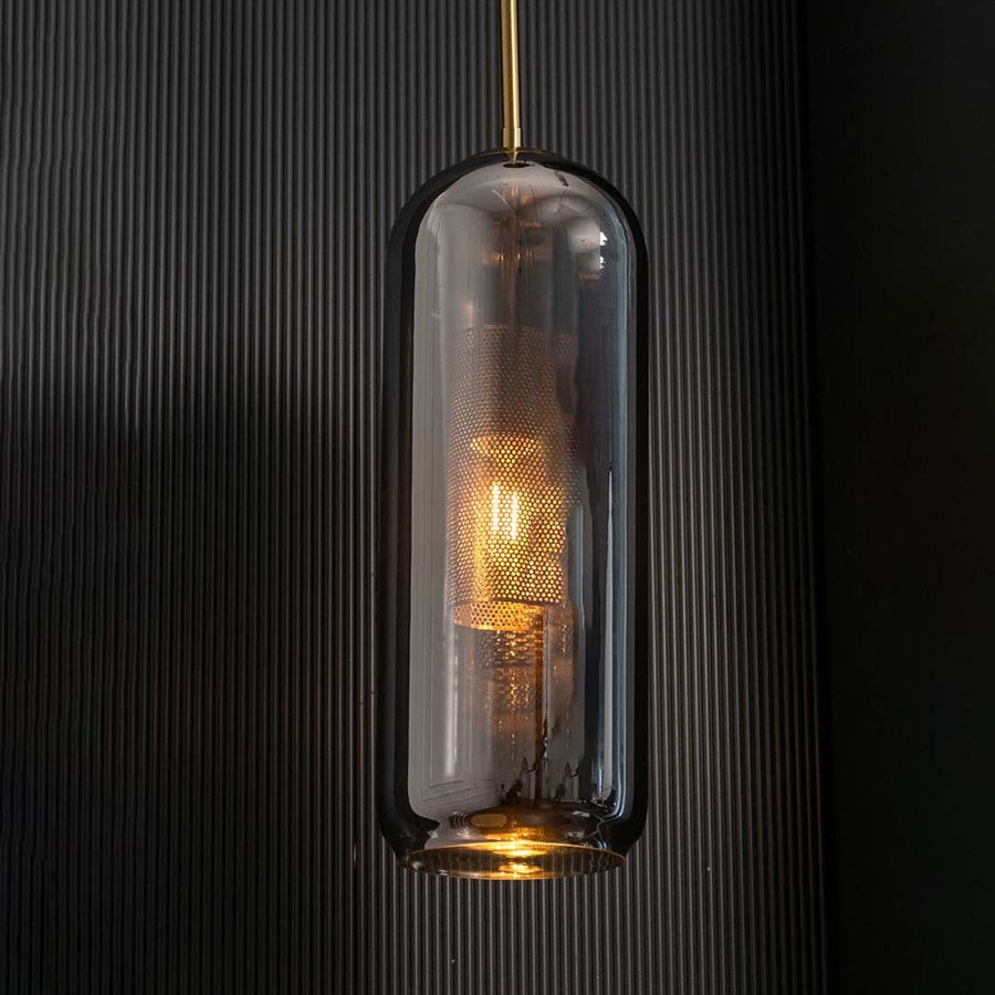 Tall smoked grey glass pendant with gold fitting and warm glowing diffuser highlighted on a dark background creating a dramatic stylish lighting accent.