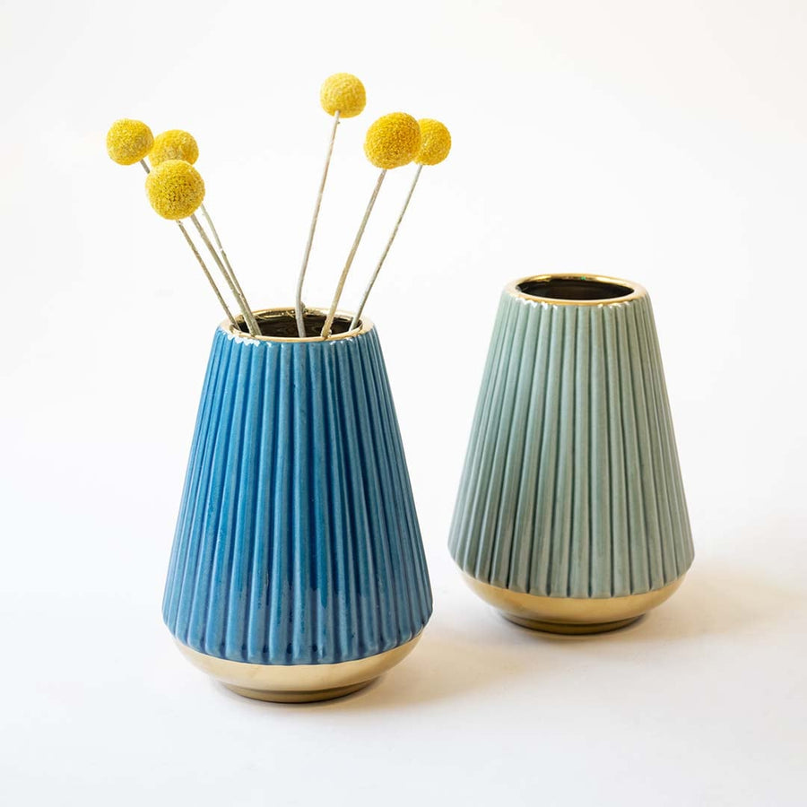 Collection of pleated ceramic vases in shades of blue and green with golden bases arranged on a nutral surface.