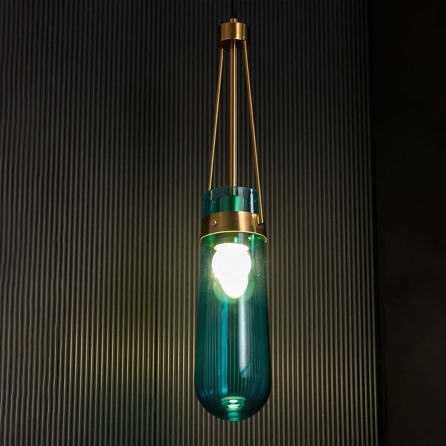 Blue glass pendant light illuminated against a black background with the light on producing a bright glow that highlights the gold frame and glass form.