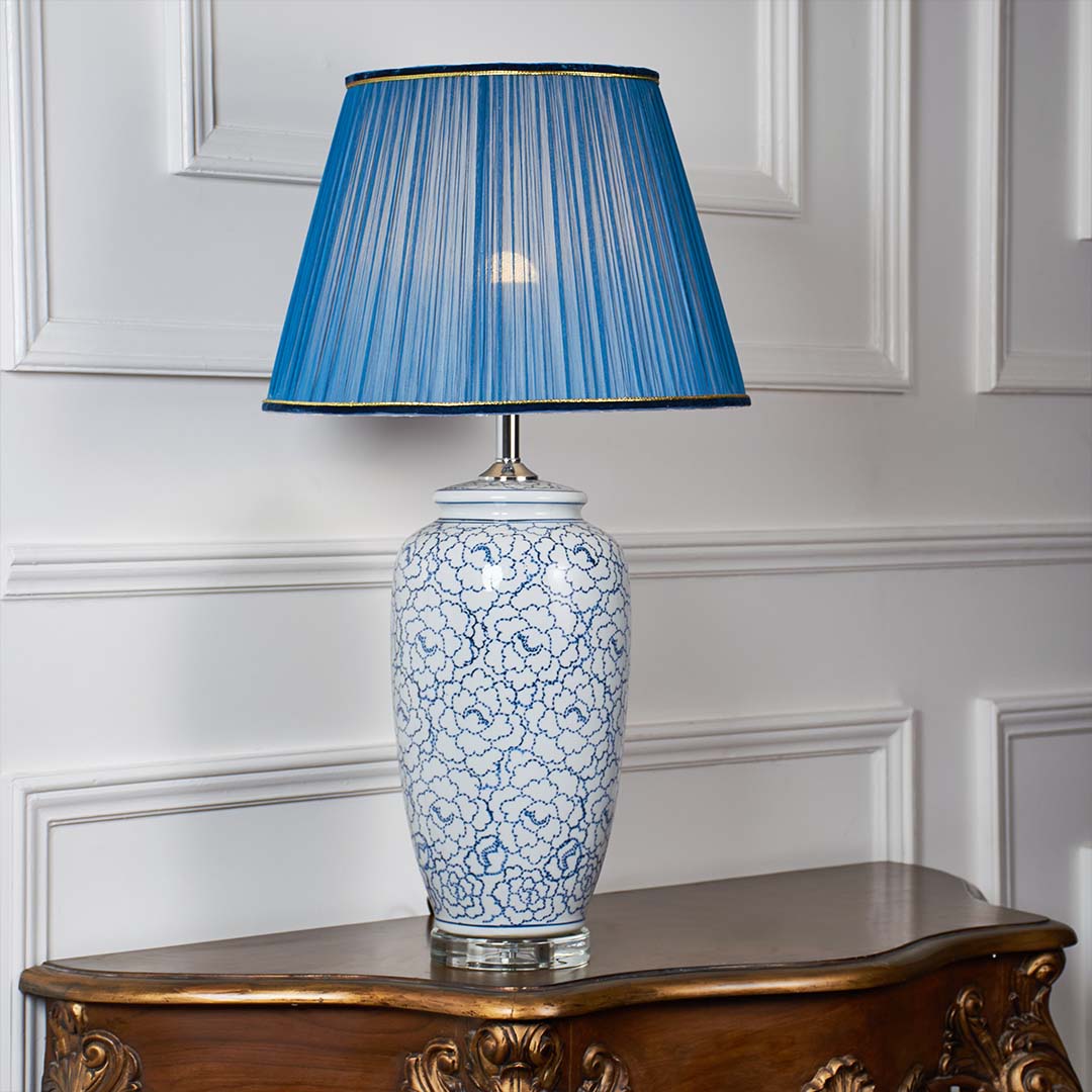 Blue floral ceramic table lamp with tall blue pleated shade glowing softly against a clean white background highlighting its detailed painted design.