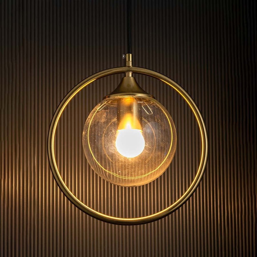 A gold ring pendant light with a clear glass globe glows warmly against a dark textured background enhancing the modern circular design with soft illumination.