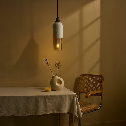 Modern white and amber glass pendant light hanging above a dining table with a cane chair and ceramic vase for a minimalist home decor look.