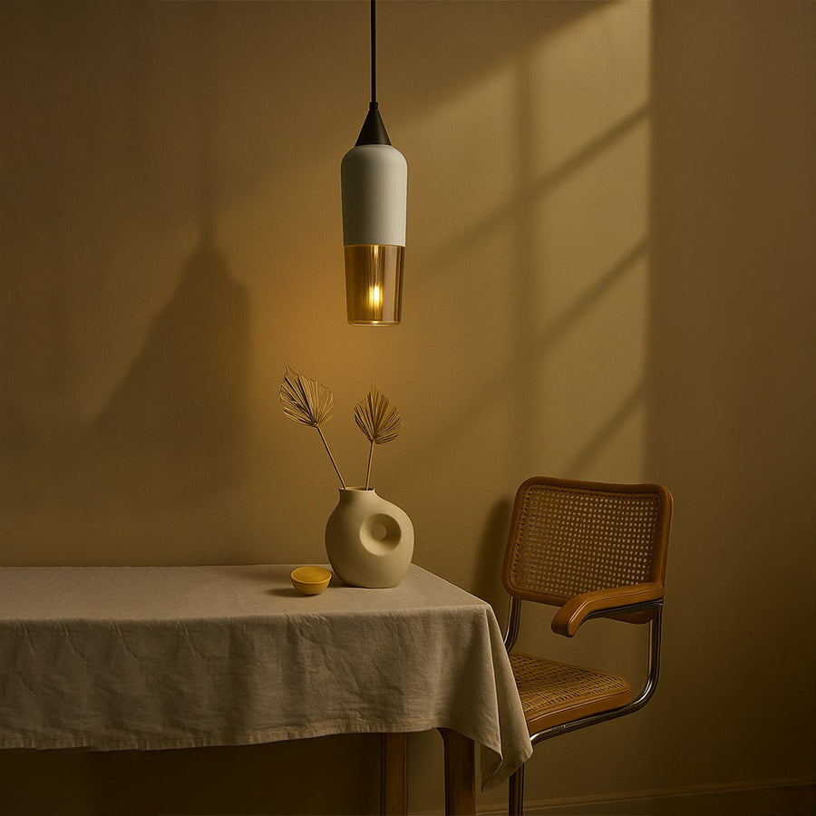 Modern white and amber glass pendant light hanging above a dining table with a cane chair and ceramic vase for a minimalist home decor look.