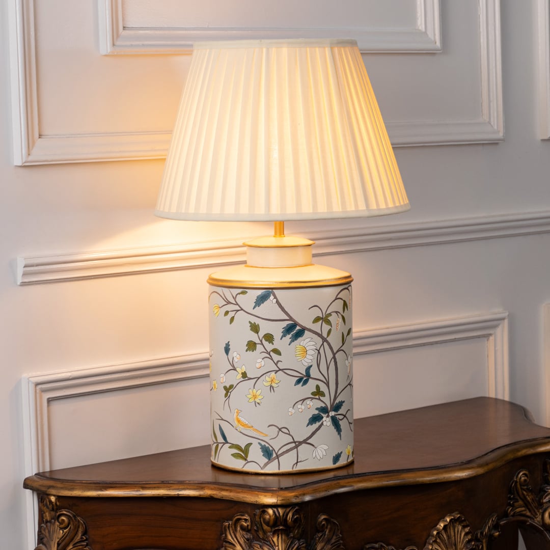 Light on white background shows ceramic table lamp with floral bird artwork and black fabric shade glowing warmly for elegant interior lighting.