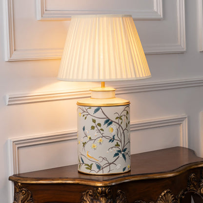 Light on white background shows ceramic table lamp with floral bird artwork and black fabric shade glowing warmly for elegant interior lighting.