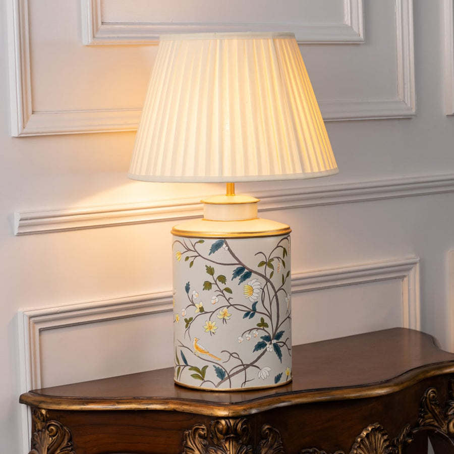 Light on white background shows ceramic table lamp with floral bird artwork and black fabric shade glowing warmly for elegant interior lighting.