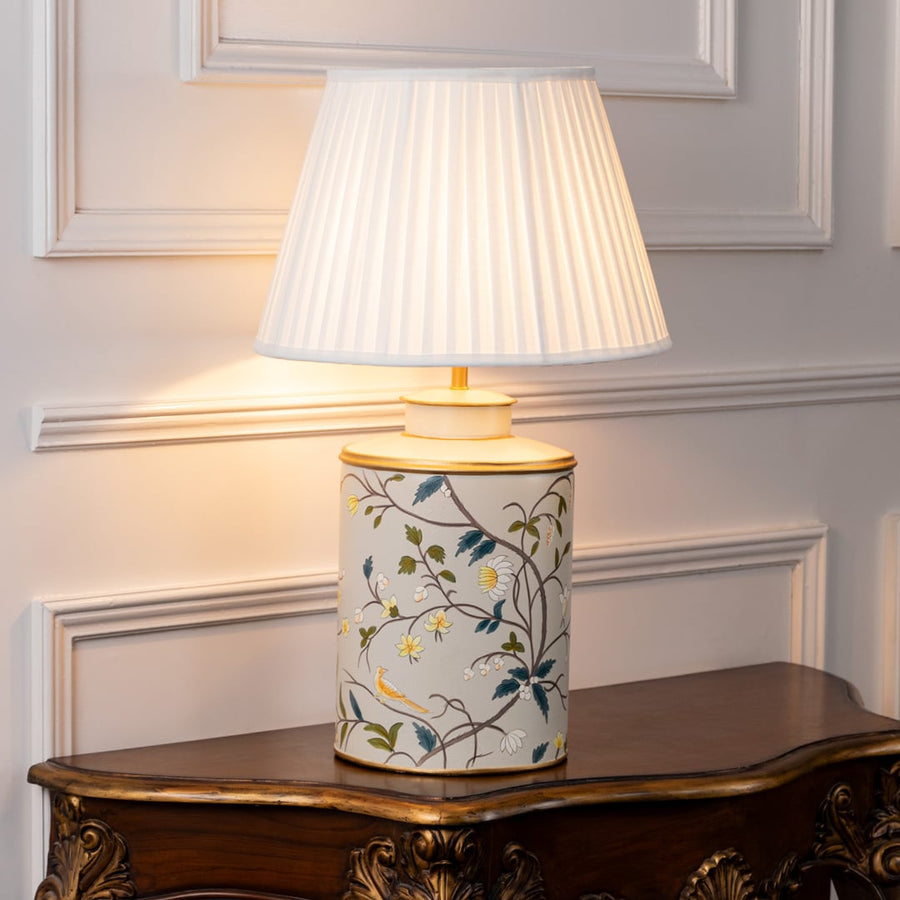 Light on white background shows ceramic table lamp with floral bird artwork and black fabric shade glowing warmly for interior lighting.