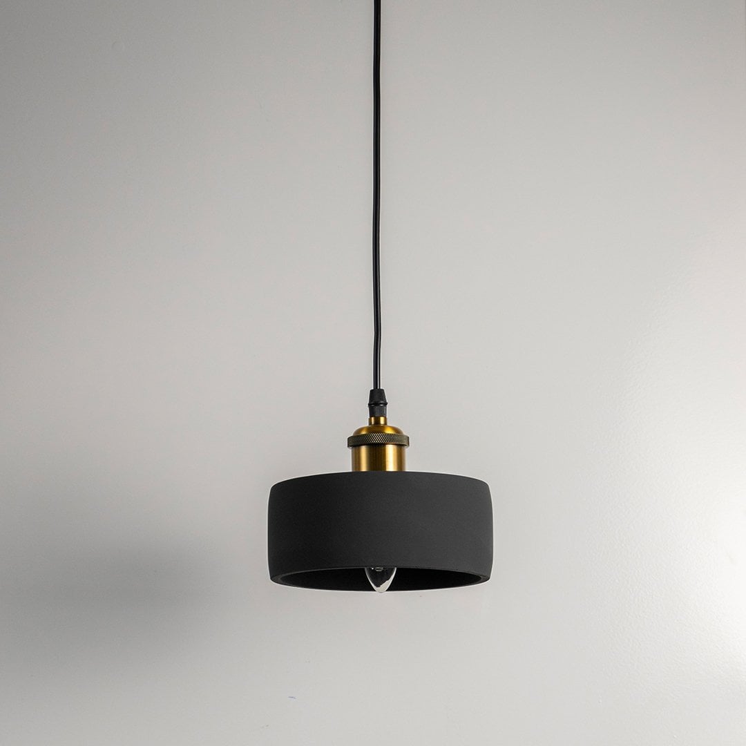 Black concrete pendant light turned off with brass top and visible bulb against white wall in modern setting.