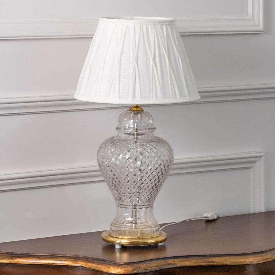 Transparent glass table lamp with gold accents and white pleated shade displayed on white background perfect for classic bedroom or side table styling.