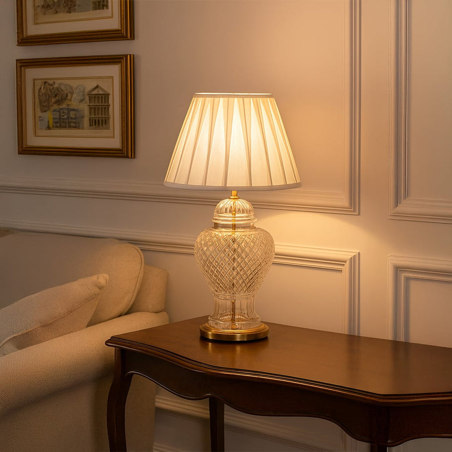 Elegant glass table lamp with gold base and white pleated shade glowing warmly on wooden console adding luxury and sophistication to living room decor.