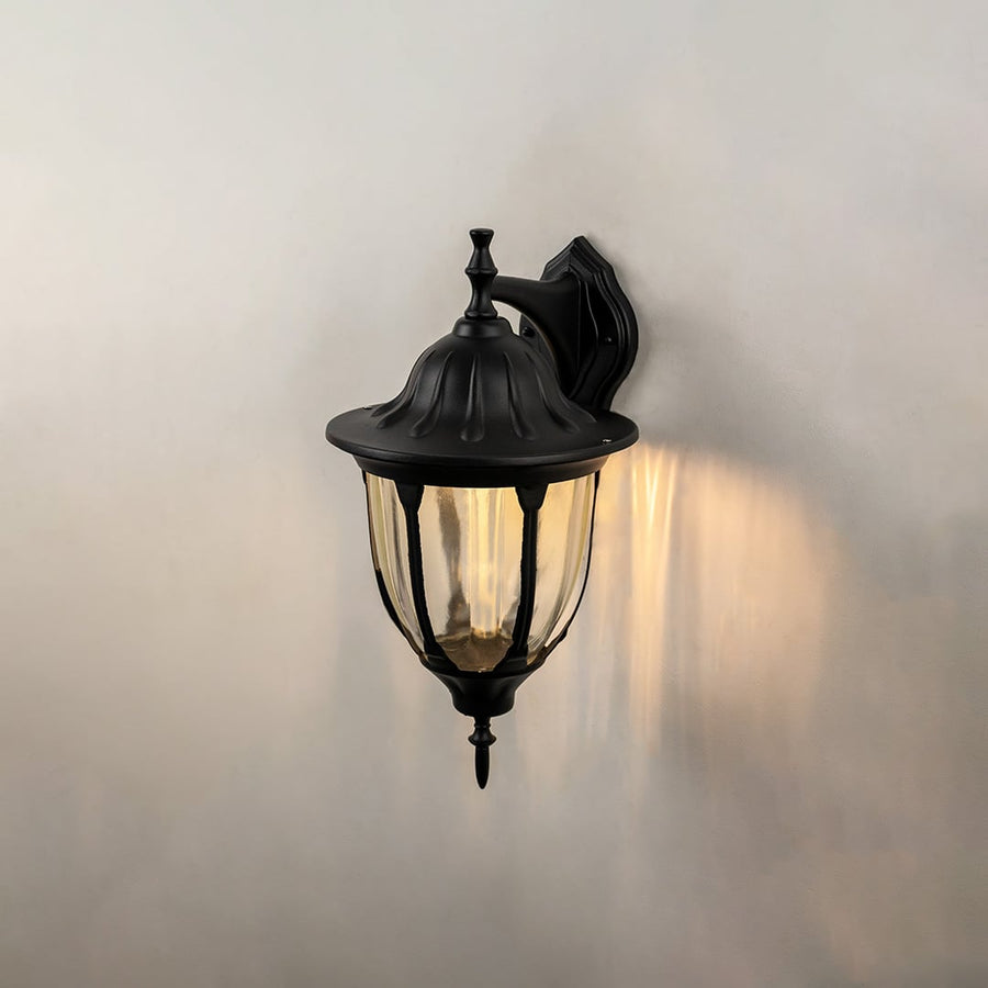Black downward outdoor wall lantern glowing on black background highlighting traditional coach style exterior lighting warm glow.