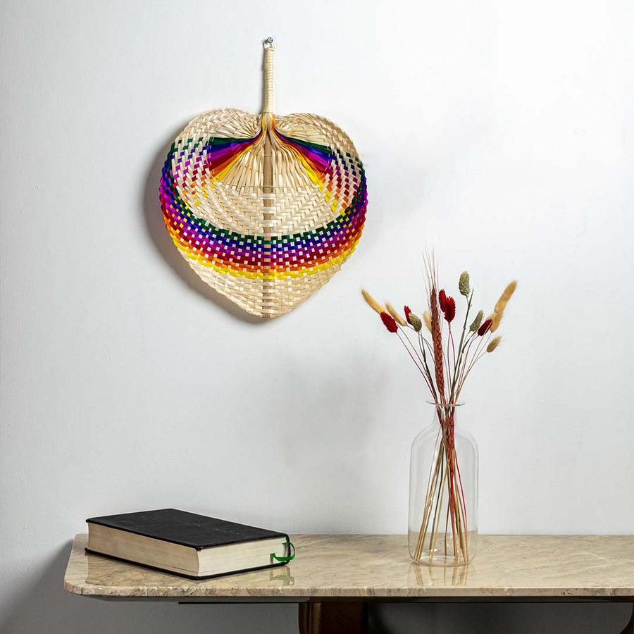 Rainbow bamboo hand fan hanging on wall creating colorful tropical bohemian accent in modern minimalist interior design space.