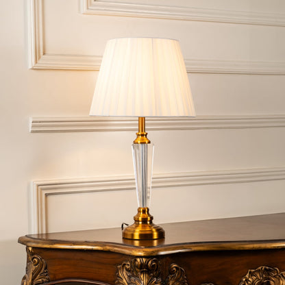 Crystal and gold table lamp with pleated shade turned on against white background perfect for bedside table lamp.