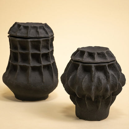Two black sculptural vases with unique grid and wave textures displayed side by side on a warm neutral background.