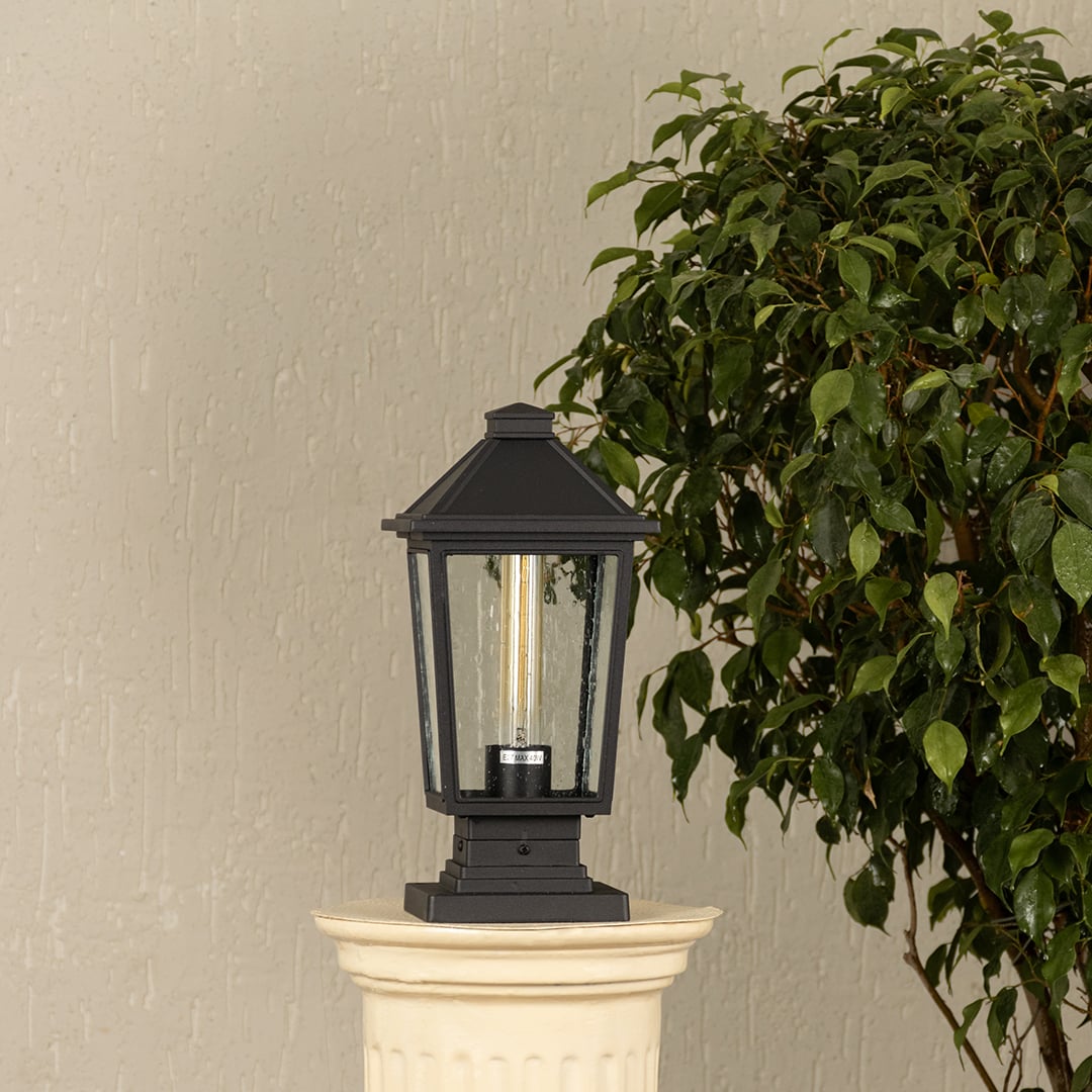 Black outdoor pillar light with clear glass panels placed by a large green plant Lifestyle image patio decor.
