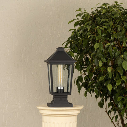 Black outdoor pillar light with clear glass panels placed by a large green plant Lifestyle image patio decor.