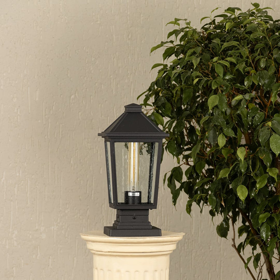 Black outdoor pillar light with clear glass panels placed by a large green plant Lifestyle image patio decor.