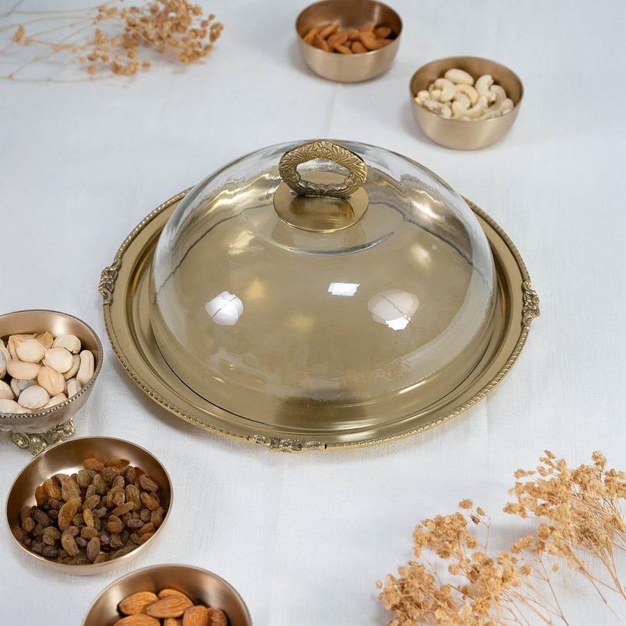 Main product image of transparent glass dome with golden handle placed on antique-style round metal tray.