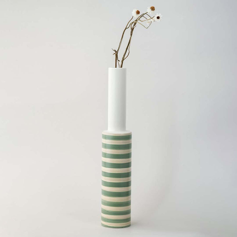 A long neck striped vase captured in bright natural light on a simple white platform showcasing its sleek artistic shape and soft green tones in a modern lifestyle scene.